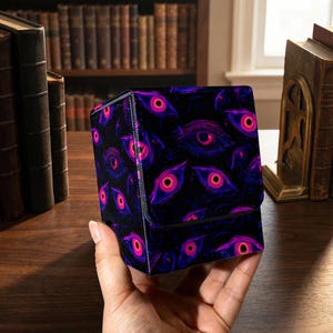 Peut inclure: Une boîte de rangement noire, en forme de cube, avec un motif répété d'yeux stylisés dans des tons de rose et de violet. La boîte est tenue dans une main, avec un fond d'étagères et de livres anciens.