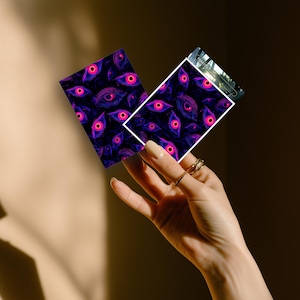 Może przedstawiać: Dwie prostokątne karty z powtarzającym się wzorem oczu w odcieniach fioletu i r&oacute;żu na ciemnoniebieskim tle. Karty są trzymane w dłoni z pierścieniami. Wz&oacute;r oka zawiera źrenicę i tęcz&oacute;wkę.