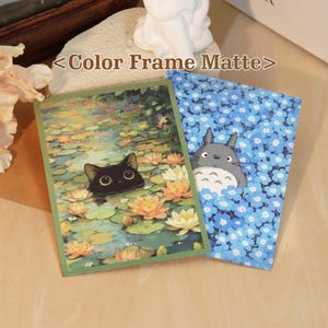 May include: Two rectangular art prints with matte frames. One print features a black cat head emerging from a pond filled with orange and yellow water lilies. The other print displays a grey creature with white markings surrounded by blue flowers.