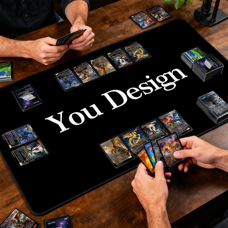 May include: A black gaming mat with the text "You Design" in white. The mat is covered with trading cards, with hands holding and arranging the cards. The cards have colorful illustrations and designs.
