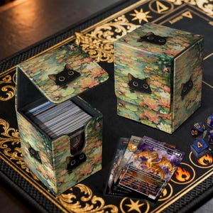 Peut inclure: Deux boîtes de rangement pour cartes avec un motif de chat et de nénuphars. Une boîte est ouverte, révélant une pile de cartes. L'autre boîte est fermée. Plusieurs cartes et dés sont sur une surface noire avec des accents dorés.