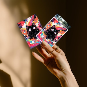 May include: Two rectangular cards featuring a black cat with large, white eyes against a colorful, swirling background of blues, pinks, and yellows. The cat's face is the focal point, with a starry design. The cards are held in a hand.