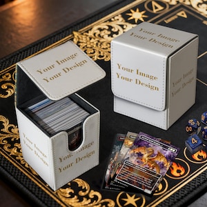 Könnte beinhalten: Zwei weiße Kartenaufbewahrungsboxen mit dem Text "Your Image Your Design" in Gold. Eine Box ist geöffnet und zeigt einen Stapel Karten. Mehrere Sammelkarten und Würfel befinden sich neben den Boxen. Die Boxen stehen auf einer schwarzen Oberfläche.