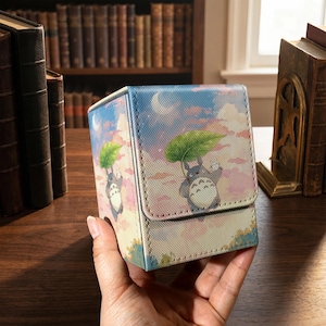Könnte beinhalten: Eine rechteckige Deck-Box mit einer aufgedruckten Illustration eines grauen, runden Wesens, das ein großes grünes Blatt als Regenschirm vor einem pastellfarbenen Himmel mit Wolken und einer Mondsichel hält. Die Box hat eine strukturierte Oberfläche.