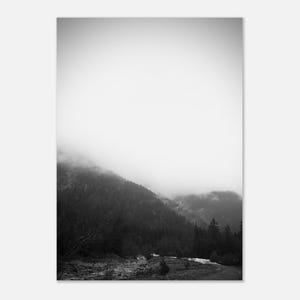 May include: Black and white landscape photograph of a mountain range shrouded in fog. The image features a dense forest covering the slopes, with a river or stream visible at the base of the mountains. The sky is a bright, overcast white.