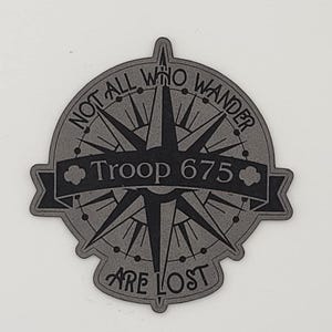 May include: A gray compass design with the text "NOT ALL WHO WANDER ARE LOST" and "Troop 675". The compass has a starburst design in the center. The text is in a bold, sans-serif font.