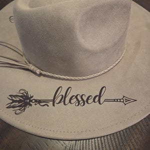 Op de afbeelding: Lichtgrijze cowboyhoed met een decoratieve touwband en een kwast. De hoed heeft het woord "blessed" in een scriptlettertype met een pijl- en veermotief. De hoed is gemaakt van een zacht materiaal.