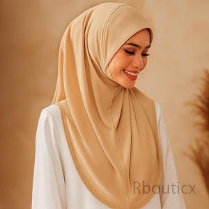 May include: A beige hijab with a matching trim, worn over a white blouse. The hijab is styled to frame the face and shoulders. The image shows a smiling person wearing the hijab. The text "Rbouticx" is visible in the bottom right corner.