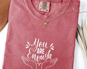 May include: A dusty rose t-shirt with the words "You are Enough" in white script. The design includes stylized hands and decorative elements. A silver necklace with a pendant is draped across the shirt.
