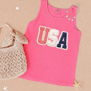 May include: A bright pink sleeveless tank top featuring the letters USA in a decorative applique style. The U is red and white striped, the S is white, and the A is navy blue with white dots. Each letter is outlined with a white scalloped trim.
