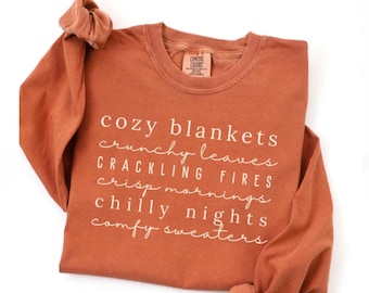May include: A burnt orange sweatshirt with the words "cozy blankets, crunchy leaves, crackling fires, crisp mornings, chilly nights, comfy sweaters" in white script. The long-sleeved shirt is folded, with the sleeves rolled up.