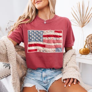 Könnte beinhalten: Ein altrosa T-Shirt mit kurzen Ärmeln und einem rechteckigen Aufdruck einer amerikanischen Flagge, die aus verschiedenen Stoffstücken zusammengesetzt ist. Das Design zeigt florale Muster, Vichy-Karos sowie rote, weiße und blaue Flächen.