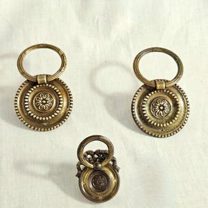 Op de afbeelding: Drie antieke messing lade grepen. Twee grotere grepen hebben een cirkelvormig ontwerp met een bloemen centrum en een ring handvat. De kleinere greep heeft een vergelijkbaar ontwerp. De grepen staan op een witte achtergrond.