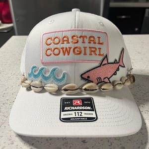 May include: White trucker hat with "COASTAL COWGIRL" text, a pink shark, and seashell accents. Features a mesh back and adjustable strap. The hat has a patch with the text "COASTAL COWGIRL" in orange letters.