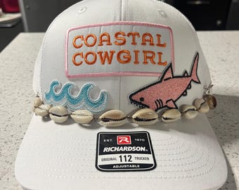 May include: White trucker hat with "COASTAL COWGIRL" text, a pink shark, and seashell accents. Features a mesh back and adjustable strap. The hat has a patch with the text "COASTAL COWGIRL" in orange letters.