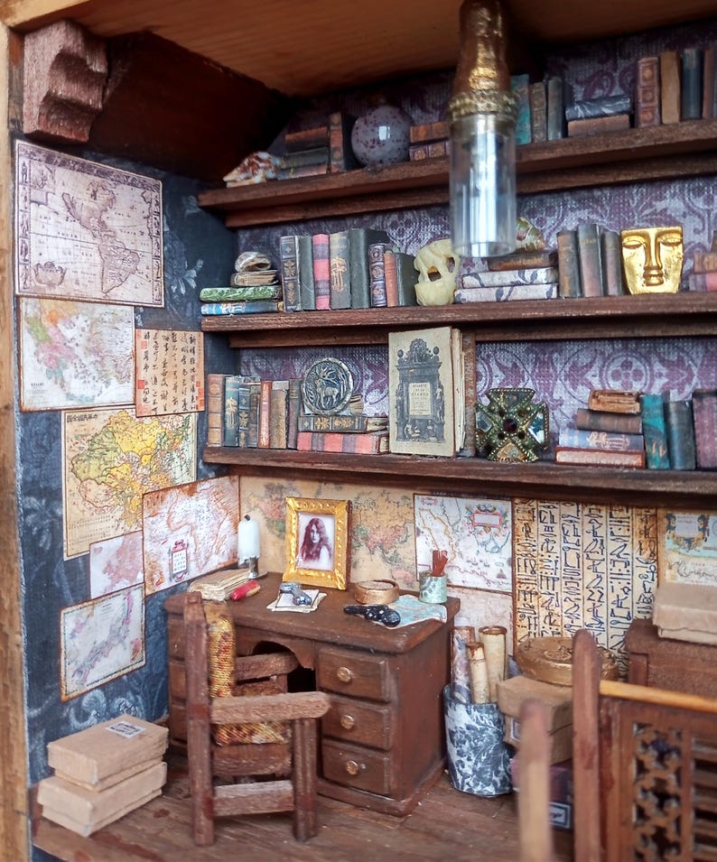 May include: Miniature library scene with a desk, chair, and bookshelves filled with tiny books. The walls are adorned with maps and framed artwork. A hanging light fixture and decorative objects complete the detailed interior.