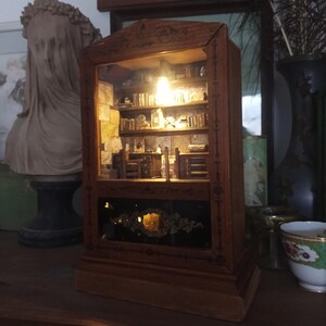 May include: A miniature wooden bookcase diorama with a warm interior light. The diorama features tiny books, furniture, and decorative elements, creating a cozy, detailed scene. The exterior is a dark wood with ornate detailing.