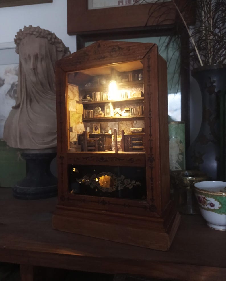 May include: A miniature wooden bookcase diorama with a warm interior light. The diorama features tiny books, furniture, and decorative elements, creating a cozy, detailed scene. The exterior is a dark wood with ornate detailing.