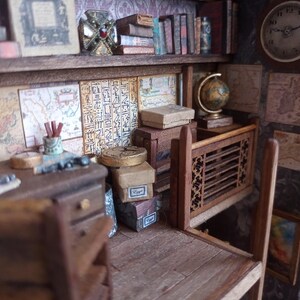 May include: Miniature library scene with a desk, shelves, books, and a globe. The shelves are filled with books of various sizes and colors. A small desk with drawers and a chair is in the foreground. The walls are decorated with maps and hieroglyphs.