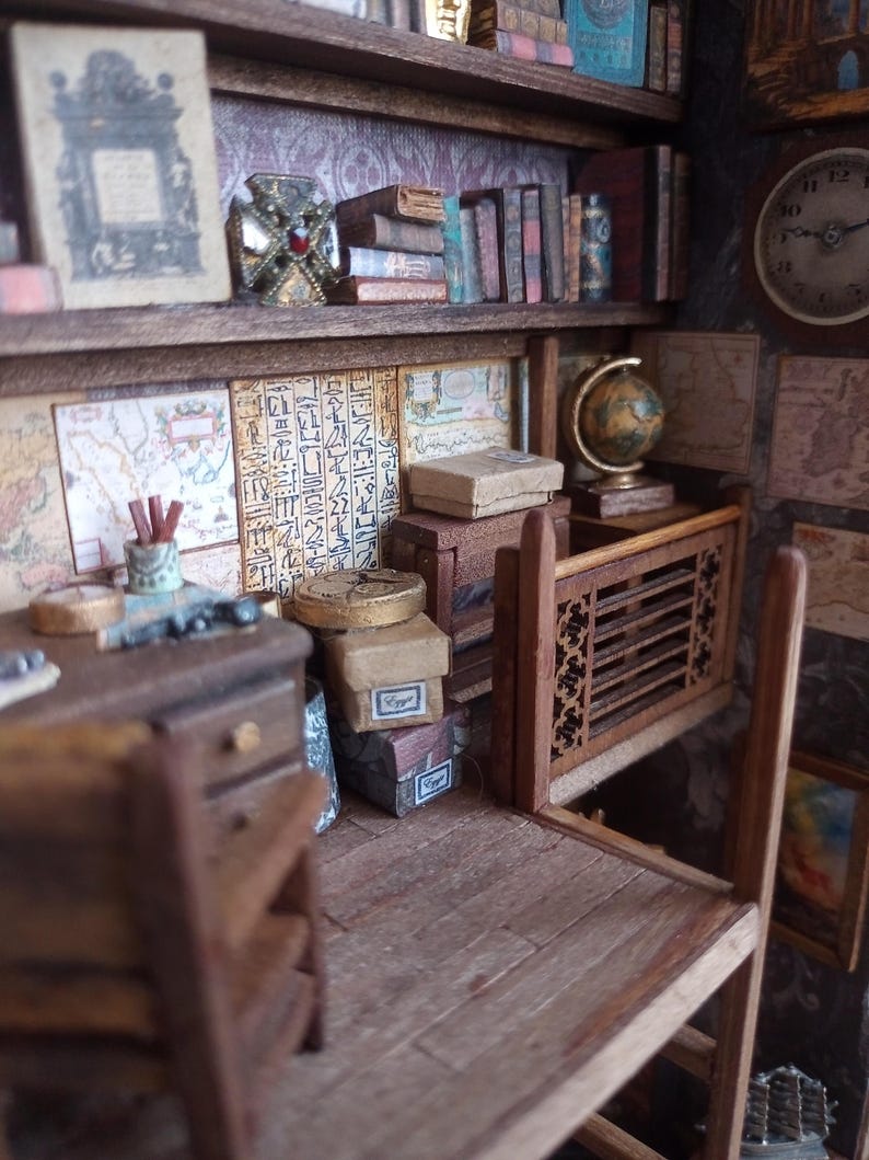May include: Miniature library scene with a desk, shelves, books, and a globe. The shelves are filled with books of various sizes and colors. A small desk with drawers and a chair is in the foreground. The walls are decorated with maps and hieroglyphs.
