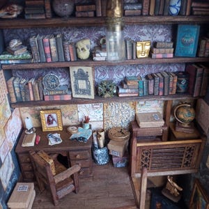 May include: Miniature library scene with wooden shelves filled with tiny books, globes, and decorative objects. A small desk with a chair, lamp, and framed picture is also present. The scene is detailed and appears to be a dollhouse or model.