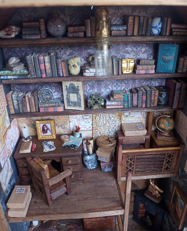 May include: Miniature library scene with wooden shelves filled with tiny books, globes, and decorative objects. A small desk with a chair, lamp, and framed picture is also present. The scene is detailed and appears to be a dollhouse or model.
