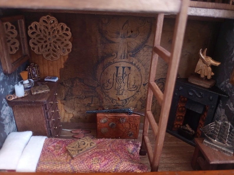 May include: Miniature diorama of a room with a bed, dresser, and fireplace. The walls are decorated with a map-like design and a large emblem. A small wooden ladder leads to a second level. A decorative piece hangs on the wall.