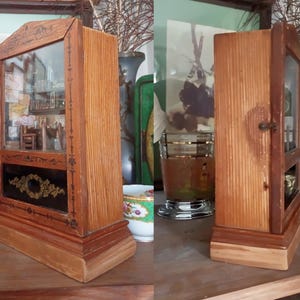 May include: A vintage wooden display cabinet with a glass front, showcasing a miniature interior scene. The cabinet has a rectangular base and a decorative black panel with gold accents. The wood has a warm, natural finish.