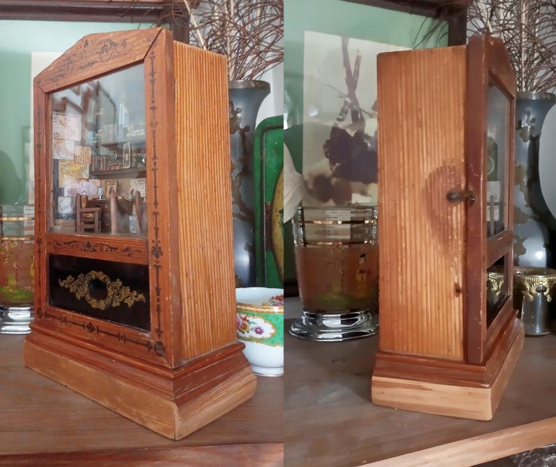 May include: A vintage wooden display cabinet with a glass front, showcasing a miniature interior scene. The cabinet has a rectangular base and a decorative black panel with gold accents. The wood has a warm, natural finish.