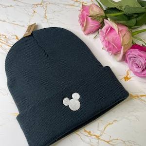 May include: Dark grey beanie hat featuring a silver Mickey Mouse head emblem. The hat is made of a ribbed knit fabric. A small tag is attached. Pink roses are in the background.