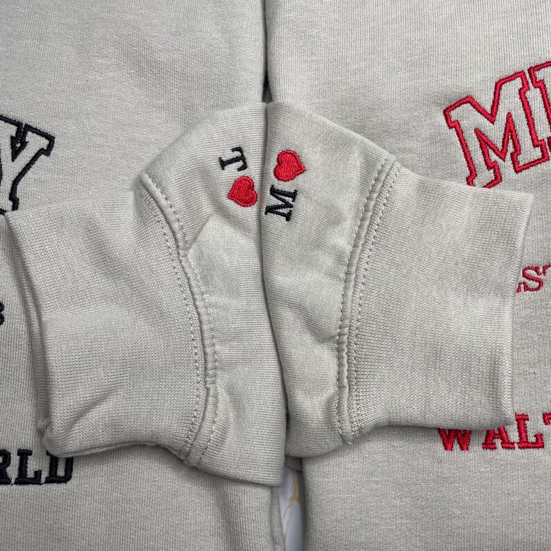 May include: Close-up of light gray sweatshirts with embroidered details. One sleeve displays "TM" with a red heart, while another shows "M" in red. Other parts of the sweatshirts have red and black lettering.