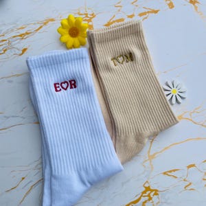 May include: Two pairs of ribbed crew socks. One pair is white with "E♡R" embroidered in red, and the other is tan with "T♡M" embroidered in gold. A yellow flower and a white daisy are also in the image.