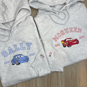 May include: Two light grey hoodies with embroidered designs. One features "SALLY" in blue with a blue car and "EST. 2020." The other displays "MCQUEEN" in red with a red car and "EST. 2020." Both have heart and initial embroidery on the sleeves.
