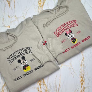 May include: Two beige hoodies with embroidered designs. One features "MICKEY" in black, a cartoon Mickey Mouse, and "WALT DISNEY WORLD". The other says "MINNIE" in red, a Minnie Mouse, and "WALT DISNEY WORLD".