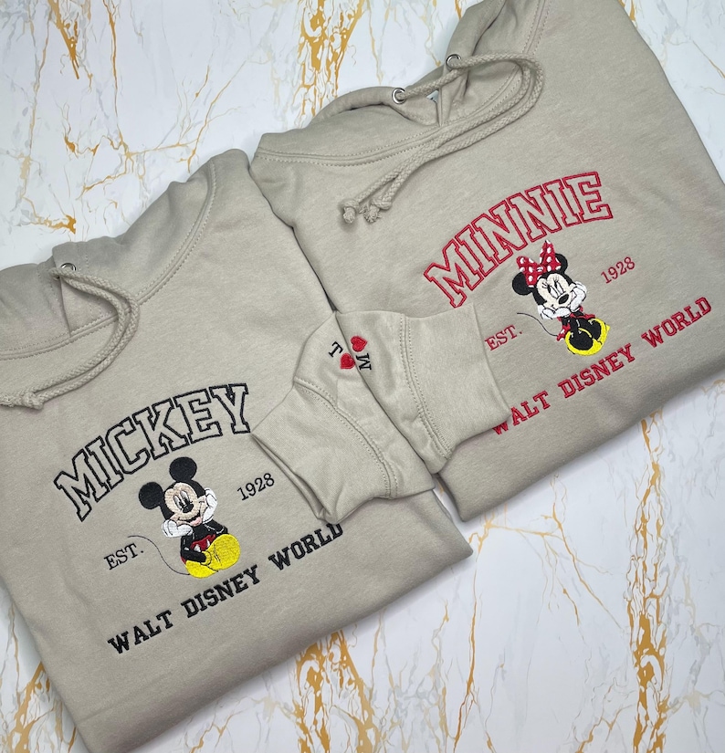 May include: Two beige hoodies with embroidered designs. One features "MICKEY" in black, a cartoon Mickey Mouse, and "WALT DISNEY WORLD". The other says "MINNIE" in red, a Minnie Mouse, and "WALT DISNEY WORLD".