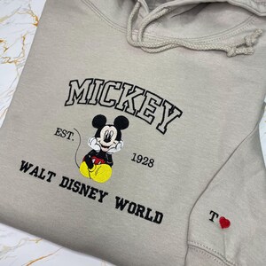 May include: Beige hoodie featuring an embroidered Mickey Mouse design. The design includes the word "MICKEY" in black, the text "EST. 1928", and "WALT DISNEY WORLD". A small red heart and the letter "T" are embroidered on the sleeve.