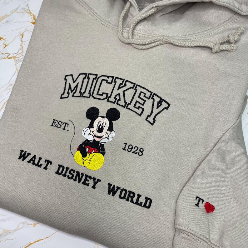 May include: Beige hoodie featuring an embroidered Mickey Mouse design. The design includes the word "MICKEY" in black, the text "EST. 1928", and "WALT DISNEY WORLD". A small red heart and the letter "T" are embroidered on the sleeve.