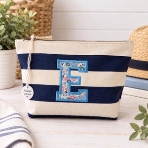 May include: A zipped pouch featuring horizontal navy blue and cream stripes with a large letter E appliqued in the centre. The letter E has a floral pattern in shades of pink, blue, and green with a light blue border. A round tag reads Handmade with love.