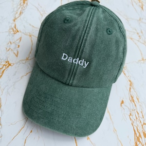 May include: A forest green baseball cap with a washed, vintage-style texture. The front panel features the word Daddy embroidered in white thread. The cap has a curved brim, stitched eyelets for ventilation, and a structured six-panel design.