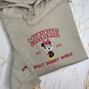 May include: A tan hoodie featuring an embroidered Minnie Mouse design. The design includes the word "MINNIE" in red, the character Minnie Mouse, and the text "EST. 1928 WALT DISNEY WORLD". The left sleeve has a small embroidered heart and the letter "M".