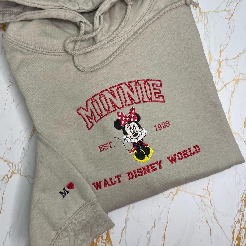 May include: A tan hoodie featuring an embroidered Minnie Mouse design. The design includes the word "MINNIE" in red, the character Minnie Mouse, and the text "EST. 1928 WALT DISNEY WORLD". The left sleeve has a small embroidered heart and the letter "M".