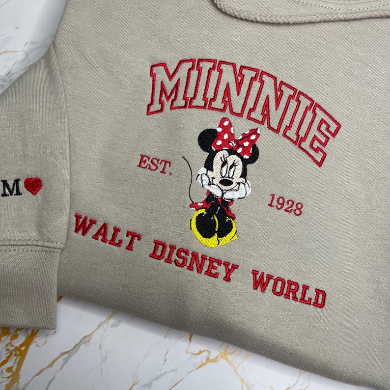 May include: Beige sweatshirt with embroidered Minnie Mouse design. The design includes the name "MINNIE" in red, the text "EST. 1928", and "WALT DISNEY WORLD". The left sleeve has the letter "M" and a red heart.