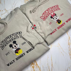May include: Two beige hoodies with embroidered designs. One features "Mickey" above a cartoon image and "Walt Disney World" below. The other displays "Minnie" with a cartoon image and "Walt Disney World". Both have the year "1928".