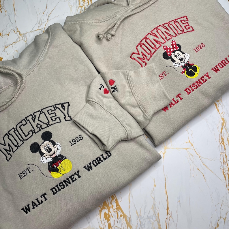 May include: Two beige hoodies with embroidered designs. One features "Mickey" above a cartoon image and "Walt Disney World" below. The other displays "Minnie" with a cartoon image and "Walt Disney World". Both have the year "1928".