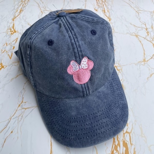 May include: A denim blue baseball cap featuring a pink Minnie Mouse bow design. The cap has a curved peak, a fabric strap, and a top button. The Minnie Mouse bow is embroidered with a pink outline and white and silver details.