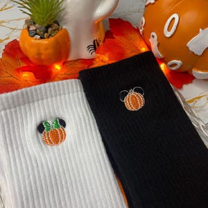 May include: Two ribbed socks, one white and one black, each featuring an embroidered pumpkin design. The pumpkin has a Minnie Mouse-inspired bow. The socks are displayed with Halloween decorations, including a ghost figurine and a pumpkin.