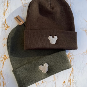 Könnte beinhalten: Zwei Strickmützen, eine dunkelbraune und eine olivgrüne, werden gezeigt. Jede Mütze hat ein silbernes Mickey Mouse Kopf-Emblem am umgeschlagenen Rand. Ein kleines Etikett der Marke "Beachfield" ist sichtbar.