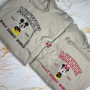 May include: Two beige hoodies with embroidered designs. One features "MICKEY" in black, a Mickey Mouse graphic, and "WALT DISNEY WORLD" in black. The other has "MINNIE" in red, a Minnie Mouse graphic, and "WALT DISNEY WORLD" in red.