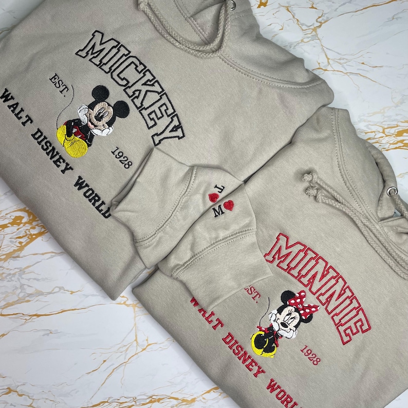 May include: Two beige hoodies with embroidered designs. One features "MICKEY" in black, a Mickey Mouse graphic, and "WALT DISNEY WORLD" in black. The other has "MINNIE" in red, a Minnie Mouse graphic, and "WALT DISNEY WORLD" in red.