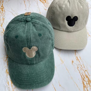 May include: Two baseball caps, one in a washed forest green with a tan embroidered mouse head silhouette and one in a light beige with a black embroidered mouse head silhouette. Both caps feature a six-panel construction with a curved brim.